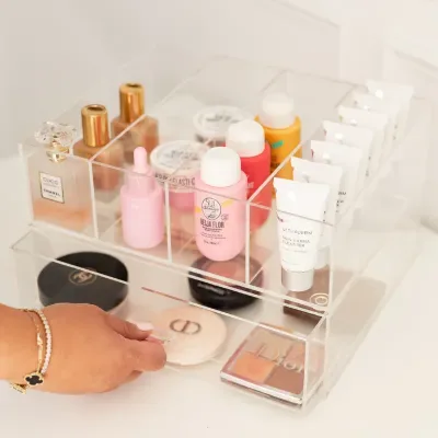 A clear acrylic drawer unit with a hand pulling the lower drawer out, holding cosmetic bottles
