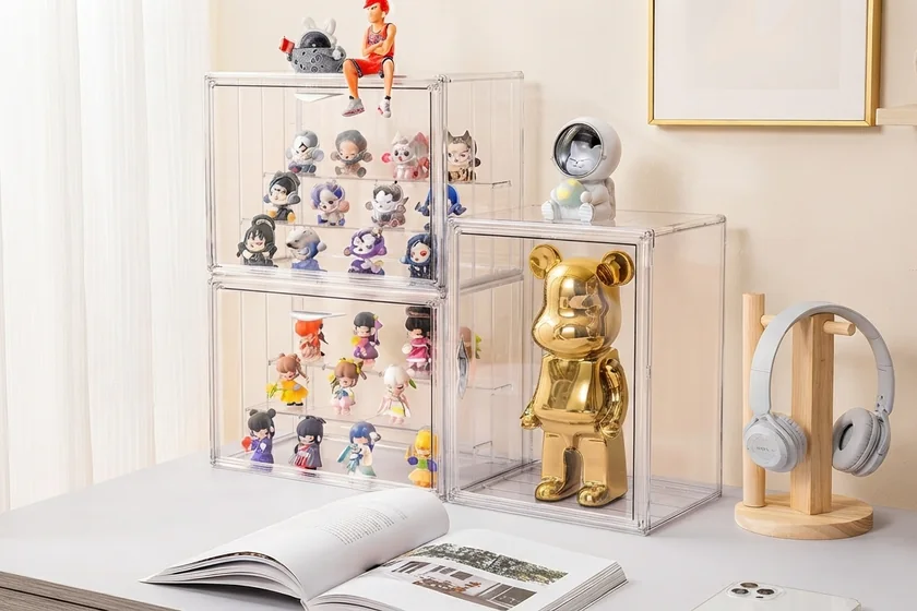 A white shelf of clear acrylic cases holding figurines, beside a teddy bear and a handbag