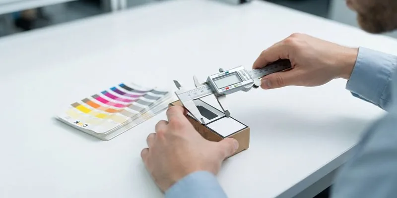 A hand using a digital caliper to measure a white vape carton beside a CMYK color swatch fan