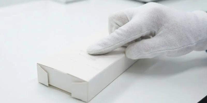 A gloved hand pressing a white folding carton on a clean inspection bench