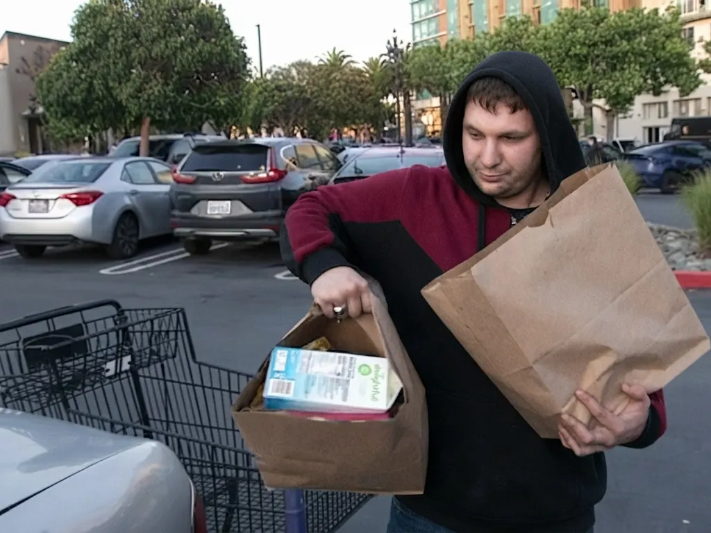 A man carrying a paper bag through a car park beside a trolley