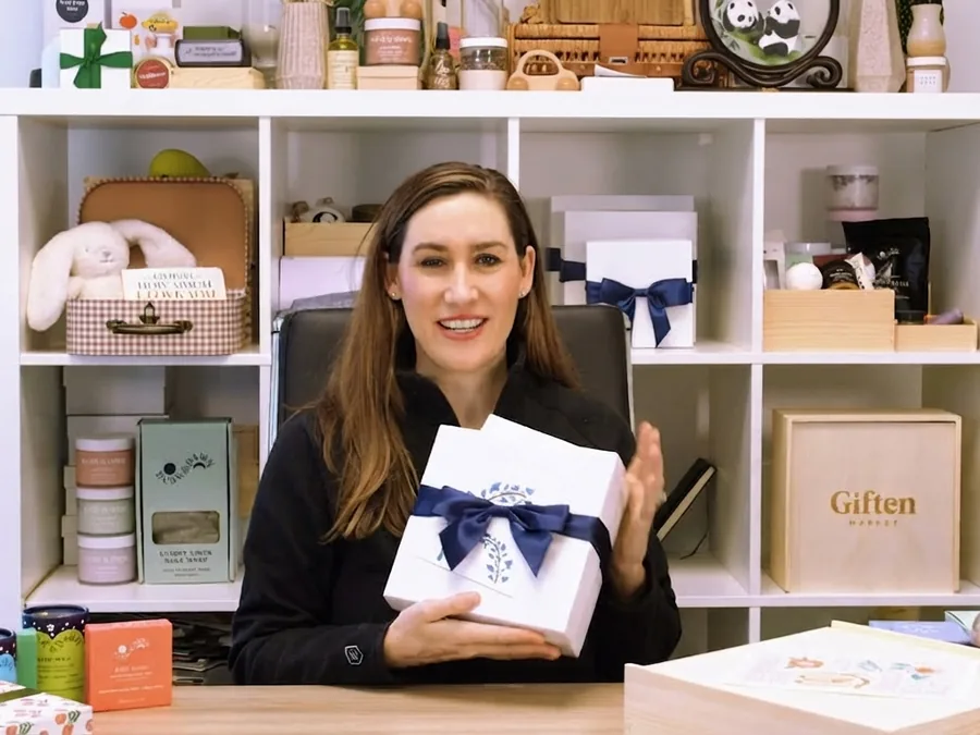 A shop owner holding a white ribboned rigid box in front of shelves of packaged goods