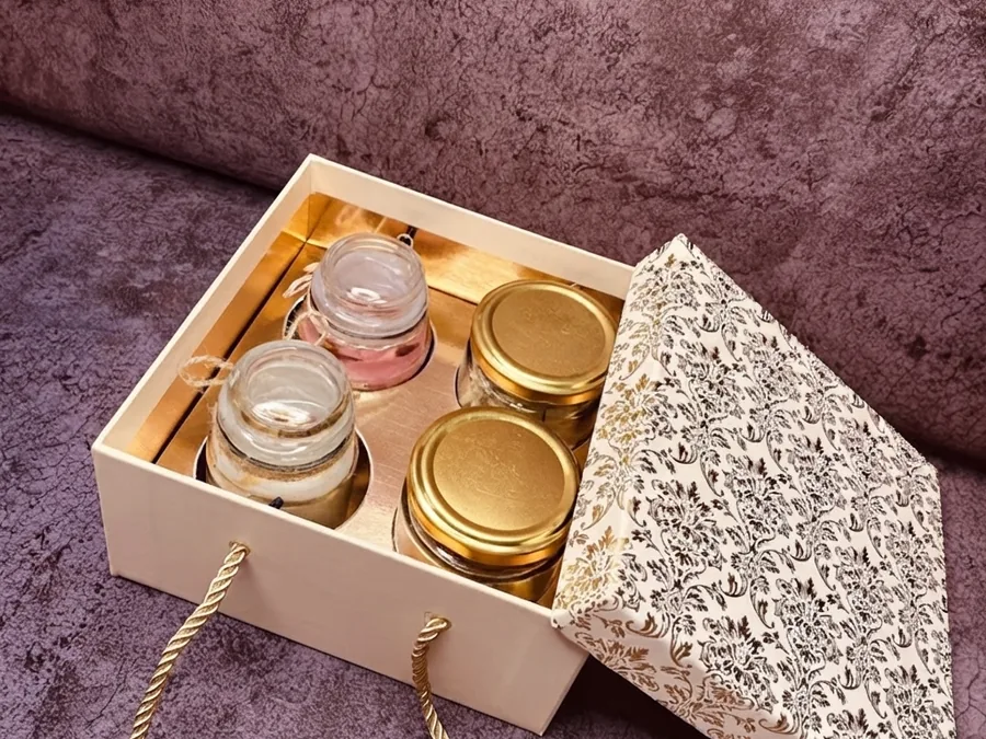 An open rigid gift box holding gold cosmetic jars, with a chain handle